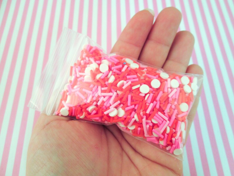 May include: A clear plastic bag filled with pink, red, and white sprinkles. The sprinkles are a mix of small round sprinkles and long, thin sprinkles.