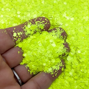 Op de afbeelding: Close-up van neongele glitter in verschillende vormen en maten. De glitter wordt in een hand gehouden, waarbij de textuur en kleur worden getoond. De glitter is een helder, opvallend materiaal.