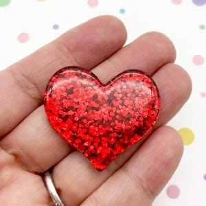 May include: A heart-shaped object with a smooth, glossy finish filled with vibrant red glitter. The heart features a clear, transparent resin-like coating that encapsulates the dense red sparkle, creating a reflective and dimensional surface.