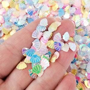 May include: Iridescent seashell-shaped confetti in pastel colors, including pink, blue, yellow, and green. The confetti is scattered, creating a shimmering effect. Ideal for crafts, scrapbooking, or party decorations.