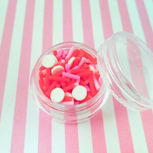 May include: A clear plastic container filled with red, pink, and white sprinkles. The container has a clear lid and is sitting on a pink and white striped surface.