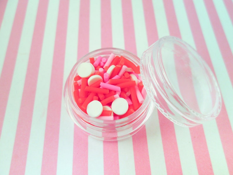 May include: A clear plastic container filled with red, pink, and white sprinkles. The container has a clear lid and is sitting on a pink and white striped surface.
