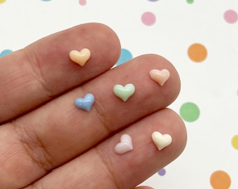 May include: A collection of six miniature heart-shaped charms in pastel colors including peach, light blue, mint green, pale pink, and cream. Each small, smooth, and glossy heart is individually placed on the surface of a finger.