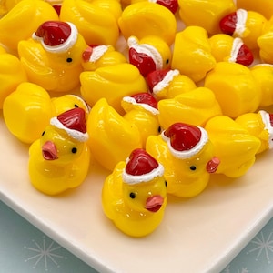Könnte beinhalten: Nahaufnahme von zahlreichen kleinen, gelben Gummienten-Figuren, von denen einige mit winzigen roten Weihnachtsmützen mit weißem Rand geschmückt sind. Die Enten sind auf einem weißen, quadratischen Teller präsentiert, was eine festliche Weihnachtsatmosphäre hervorruft. Die Enten haben schwarze Augen und rosa Schnäbel.