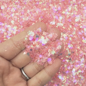 Op de afbeelding: Close-up van roze glitter met iriserende accenten. De glitter wordt in een hand gehouden, waardoor de textuur en glans zichtbaar worden. De glitter lijkt een mix van vormen en maten te zijn, wat een oogverblindend effect creëert.