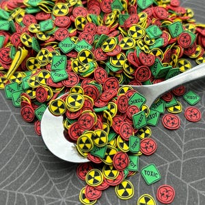 May include: A pile of colorful, round and square sprinkles with a biohazard symbol and the word "TOXIC" printed on them. The sprinkles are in a silver spoon on a black and green spiderweb background.