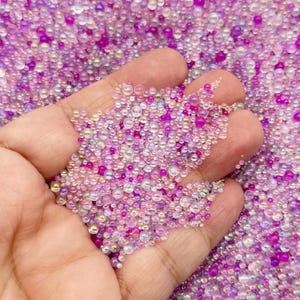 May include: A handful of small, iridescent beads in shades of pink, purple, and clear. The beads are of varying sizes and shapes, creating a textured and visually appealing display. The beads are held in an open hand, with more beads scattered in the background.