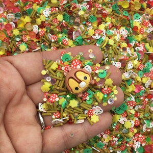 May include: A close-up of a hand holding a variety of colorful sprinkles, including green frogs, red mushrooms, yellow flowers, and brown sticks. There is also a small brown and yellow cartoon character sprinkle.