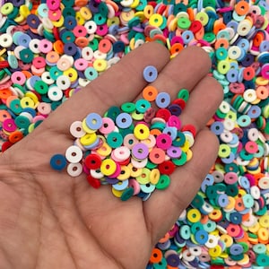Op de afbeelding: Een hand die een verscheidenheid aan kleurrijke donutvormige kralen vasthoudt. De kralen zijn gemaakt van polymer clay en zijn verkrijgbaar in verschillende kleuren, waaronder roze, blauw, groen, geel en oranje.