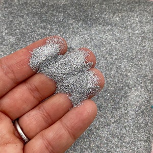 May include: A close-up of a hand holding a pile of silver glitter. The glitter is very fine and has a slight iridescent sheen.