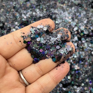May include: A close-up of a hand holding a variety of iridescent glitter in shades of black, silver, and purple.