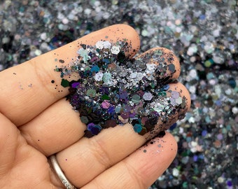 Pode incluir: Um close-up de uma mão segurando uma variedade de glitter iridescente em tons de preto, prata e roxo.