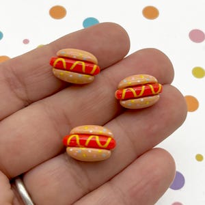 May include: Three miniature hot dog charms made of polymer clay. Each charm is a light brown bun with a red hot dog, yellow mustard, and white sprinkles.