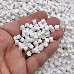 May include: A hand holding a pile of small, white, round beads. The beads are about the size of a pea and have a smooth, matte finish.