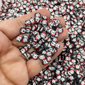 May include: A hand holding a large quantity of small, round, red and black Christmas-themed resin embellishments. Each embellishment features a white cartoon face with a black hat.