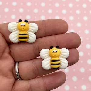 May include: Two small, yellow and black bee-shaped embellishments with white wings. The bees have a smiling face and are held in a hand against a pink background with white polka dots. These are likely craft supplies.