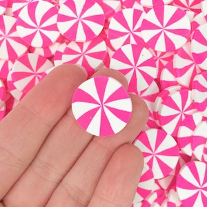 May include: A hand holding a pink and white candy-shaped embellishment. The embellishment is round with a pinwheel design. The background is filled with similar embellishments. These are likely used for crafts or decorations.
