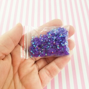 May include: A small clear plastic bag filled with iridescent purple rhinestones. The rhinestones are round and reflect multiple colors. The bag has a red zipper and is held in a hand against a pink and white striped background.