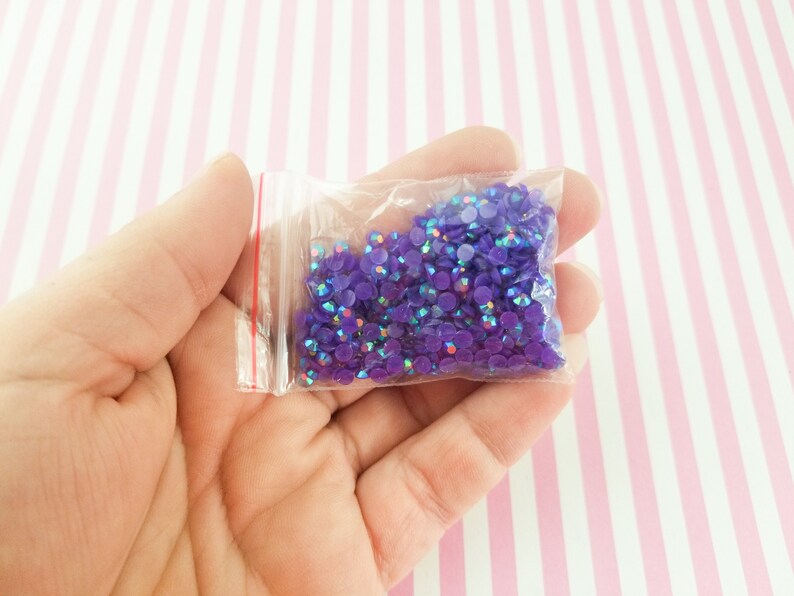May include: A small clear plastic bag filled with iridescent purple rhinestones. The rhinestones are round and reflect multiple colors. The bag has a red zipper and is held in a hand against a pink and white striped background.