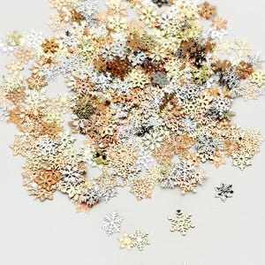 May include: A collection of metallic snowflake-shaped confetti in various colors, including gold, silver, and rose gold. The confetti is scattered across a light-colored surface, creating a festive and decorative effect. Ideal for holiday crafts and decorations.