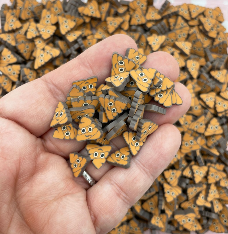 May include: A hand holding a pile of small, brown and orange, poop emoji shaped resin charms.