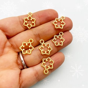 May include: Six gold-colored snowflake charms with a small loop at the top for hanging. The charms are held in a hand, and the background is a light blue color with white snowflake designs.