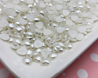May include: A white ceramic dish filled with heart-shaped beads. The beads have a pearlescent finish on one side and a flat, white surface on the other. The beads are scattered across the dish, creating a textured surface.
