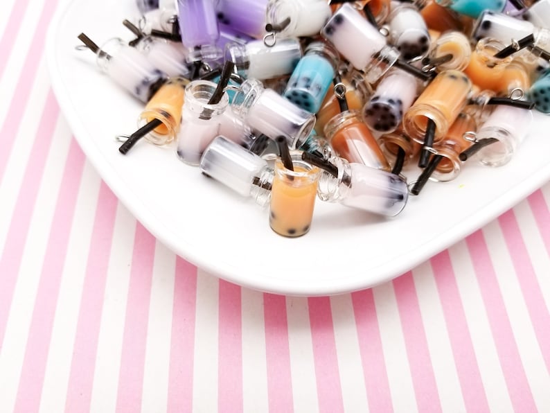 Pu&ograve; includere: Una collezione di ciondoli di bubble tea in plastica miniaturizzati colorati con cannucce nere. I ciondoli sono di vari colori, tra cui rosa, blu, viola e marrone. I ciondoli sono disposti su un piatto bianco con uno sfondo a strisce rosa e bianco.