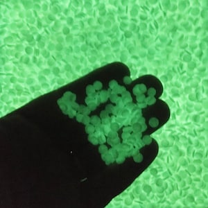 May include: A hand holds a small pile of glowing green beads, with a background of similar beads. The beads are round and emit a bright, soft green light. The hand is silhouetted against the glowing beads.