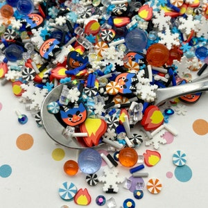 May include: A variety of colorful sprinkles and decorative elements, including blue and red cartoon faces, white snowflakes, orange and blue candy swirls, and clear and blue glass beads. The sprinkles are scattered on a white surface with colorful polka dots.
