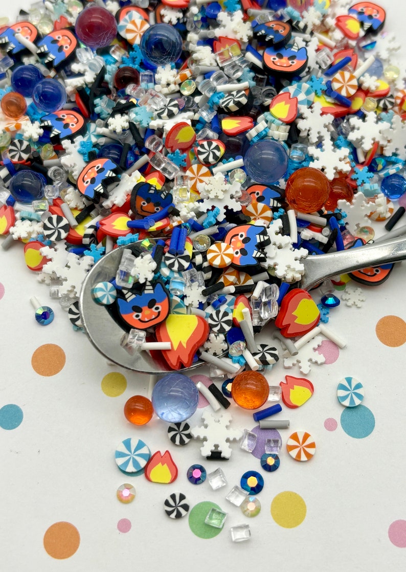 Peut inclure: Une vari&eacute;t&eacute; de vermicelles color&eacute;s et d'&eacute;l&eacute;ments d&eacute;coratifs, y compris des visages de dessins anim&eacute;s bleus et rouges, des flocons de neige blancs, des spirales de bonbons orange et bleus, et des perles de verre transparentes et bleues. Les vermicelles sont dispers&eacute;s sur une surface blanche avec des pois color&eacute;s.