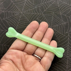 May include: A light green, bone-shaped object. The bone is held in a hand against a black background with a white spiderweb pattern. The bone is a solid color and has a smooth texture. The object is likely a Halloween decoration.