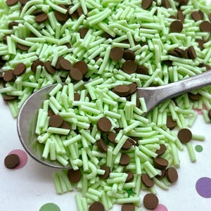 May include: Green and brown sprinkles for baking and decorating. The sprinkles are in the shape of long, thin sticks and small, round discs.