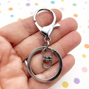 Puede incluir: Un llavero plateado con un mosquetón, un llavero y un pequeño conector metálico. El llavero está diseñado para sujetar llaves u otros objetos pequeños. El fondo es un patrón colorido de puntos.