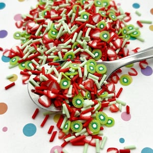 May include: A close-up of a spoon filled with red and green sprinkles. The sprinkles are shaped like strawberries and kiwi slices, and there are also small red and green sticks. The sprinkles are on a white background with colorful polka dots.