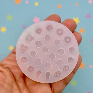 May include: A clear, circular silicone mould featuring a variety of miniature food shapes, including swirls, dots, and other designs. The mould is held in a hand against a light blue background with colourful star shapes.