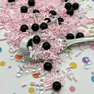 May include: Pink and clear sprinkles with black round sprinkles. The sprinkles are on a white surface with colorful polka dots.