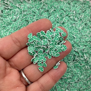 May include: A collection of small, green and white shamrock-shaped polymer clay pieces. The shamrocks have a white outline and are scattered on a background of similar shamrocks. These are likely used for crafting or decorative purposes.
