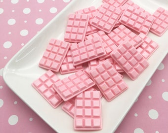 May include: A collection of small, rectangular, light pink resin charms shaped like miniature chocolate bars with a grid pattern on the surface. These items are arranged in a pile on a white, rectangular ceramic tray with rounded corners.