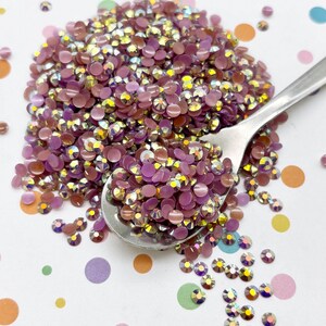 May include: A pile of iridescent, mauve-colored rhinestones, some held in a silver spoon. The rhinestones are scattered on a white surface decorated with colorful polka dots. These small, round gems sparkle.