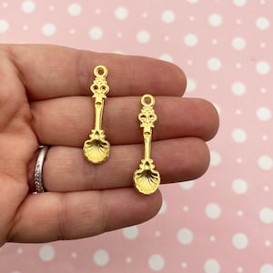 Op de afbeelding: Twee gouden metalen lepelscharmen met sierlijke details en een schelpvormige kom.