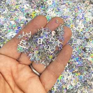 Könnte beinhalten: Eine Handvoll schimmernder silberner Buchstaben-Glitzer. Der Glitzer hat die Form von Buchstaben des Alphabets.