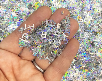 May include: A handful of iridescent silver letter-shaped glitter. The glitter is in the shape of letters of the alphabet.