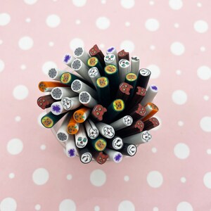 May include: A collection of polymer clay canes with Halloween-themed designs. The canes feature images of spiders, spiderwebs, jack-o'-lanterns, skulls, and ghosts. The canes are in a clear container against a pink background with white polka dots.