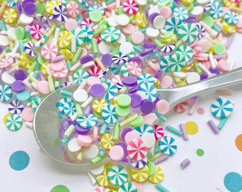 Pode incluir: Uma variedade de granulados coloridos numa colher, incluindo rosa pastel, verde, azul, roxo e amarelo. Os granulados têm a forma de pequenos círculos, bastões de caramelo e palitos longos e finos.