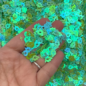 May include: A close-up of a handful of iridescent green leaf-shaped glitter. The glitter is scattered on a surface and is being held in a hand.