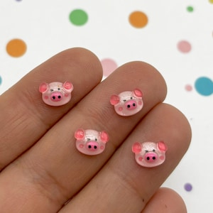 May include: Four pink pig face resin charms with black eyes and pink noses. The charms are glossy and have a slight sparkle.