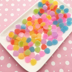May include: A white rectangular plate filled with colourful, round sweets. The sweets are in various colours, including pink, orange, yellow, green, and blue. The sweets have a sugared coating. The plate is on a pink background with white polka dots.