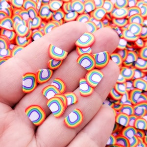 Puede incluir: Una mano sosteniendo una variedad de espolvoreos de arcilla polimérica en forma de semicírculo de color arcoíris. Los espolvoreos son perfectos para agregar un toque de fantasía a las manualidades y proyectos.