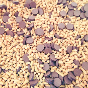 May include: Close-up of a mix of baking ingredients. The image shows a combination of small, light brown, rectangular pieces and dark brown, round chocolate chips. The ingredients are scattered across the surface.